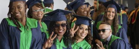 A group of Delaware Tech graduates in their graduation regalia standing in a group posed together for a photo, some are smiling and some are holding up two fingers to signify a peace sign